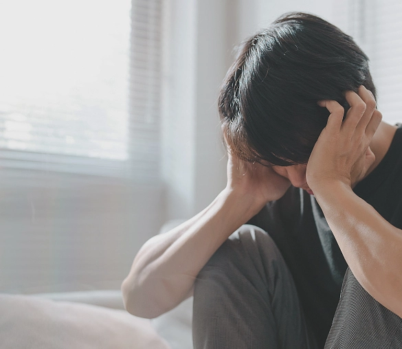 Junger Mensch sitzt zusammengekauert in einem Zimmer und hält sich die Hände an den Kopf