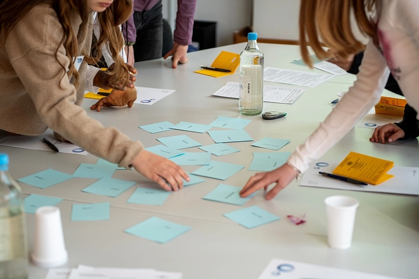Kinder stehen um einen Tisch herum und greifen nach kleinen blauen Zetteln, die auf dem Tisch verteilt liegen. Vor den Kindern liegen gelbe Impfbücher.